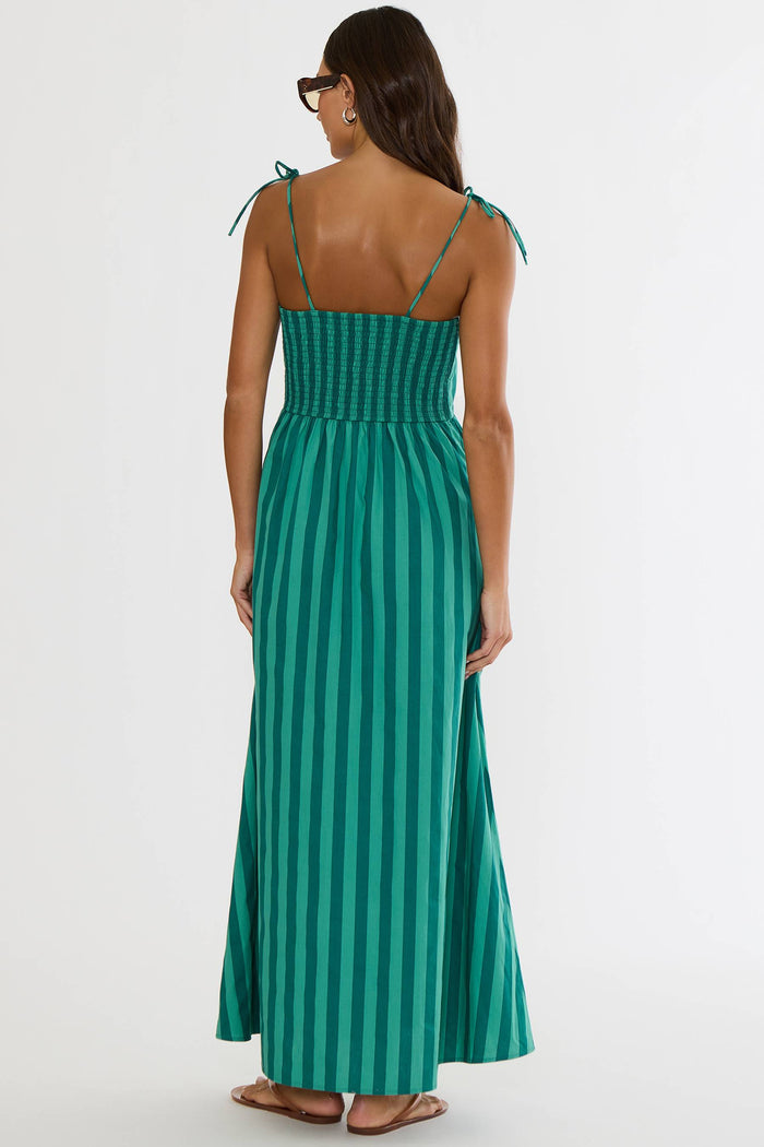 A woman shown from the back wearing a green and teal vertically striped maxi dress with thin shoulder ties, photographed against a plain light gray studio background.