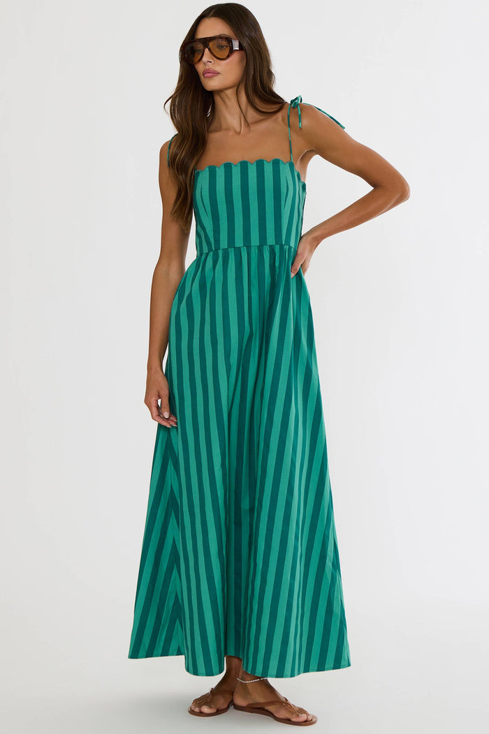 A woman modeling a green and teal vertically striped maxi dress with thin shoulder straps and side ties, paired with brown flat sandals and sunglasses, against a plain light gray studio background.