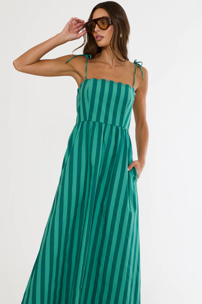 A woman modeling a green and teal vertically striped maxi dress with thin shoulder ties and side pockets, photographed against a plain white studio background.