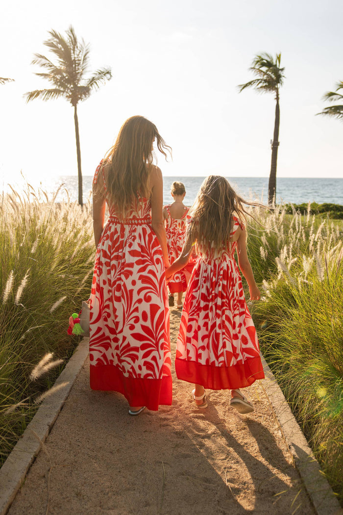 Two women and a young girl in red and white dresses walking along a path with palm trees and ocean in the background.