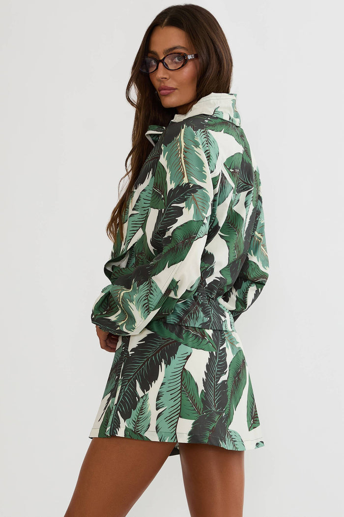 A woman wearing a green and white tropical leaf print hooded jacket and matching mini skirt, photographed against a plain light gray studio background.