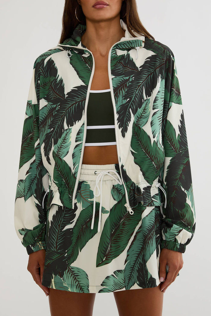 A woman wearing a matching tropical print windbreaker jacket and shorts set. The set features a bold green and black palm leaf pattern on an off-white background, styled over a black and white striped crop top.