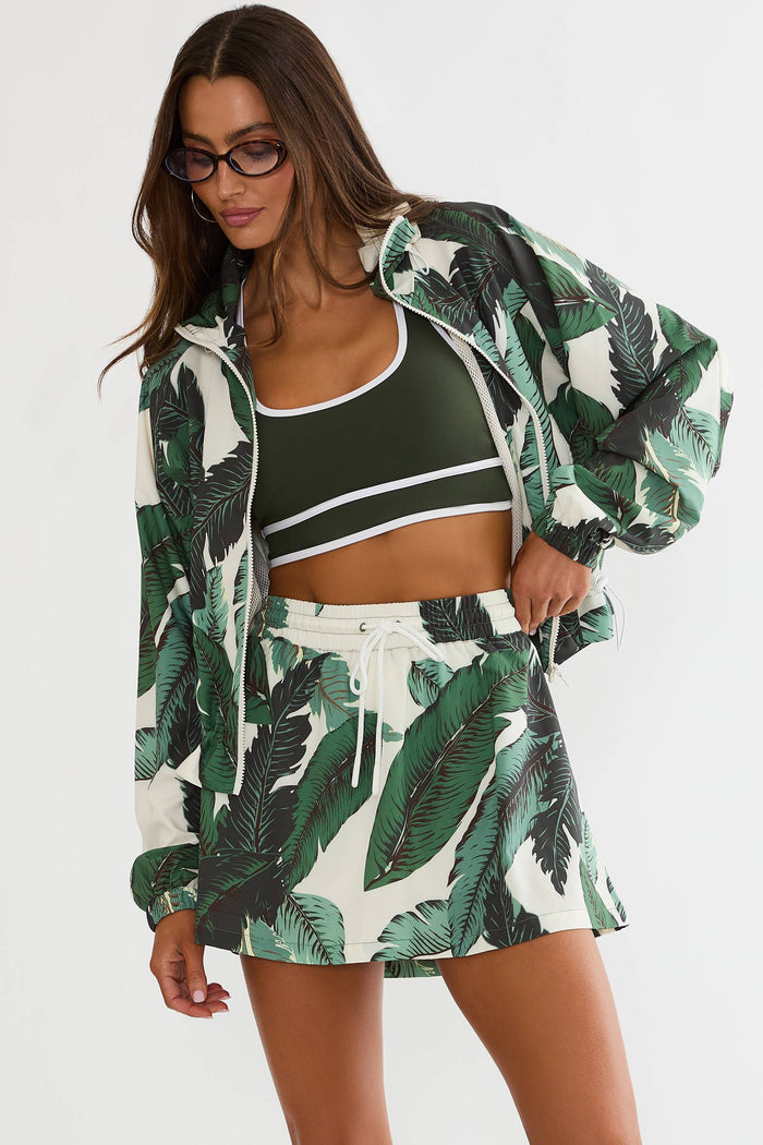 A woman modeling a matching tropical print lounge set consisting of a long-sleeve zip-up jacket and high-waisted shorts, worn over a dark green sports bra, against a plain light gray background.