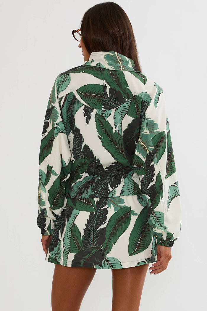 A woman shown from the back wearing a long-sleeve collared shirt dress with a green tropical leaf print on an off-white background, set against a plain light gray studio backdrop.