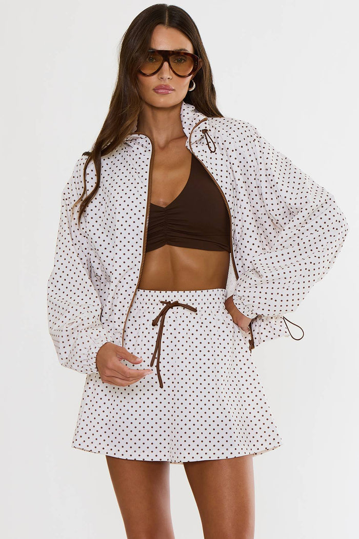 A model wearing a white long-sleeve zip-up jacket and matching mini skirt set with small brown polka dots, styled over a dark brown crop top and accessorized with sunglasses.