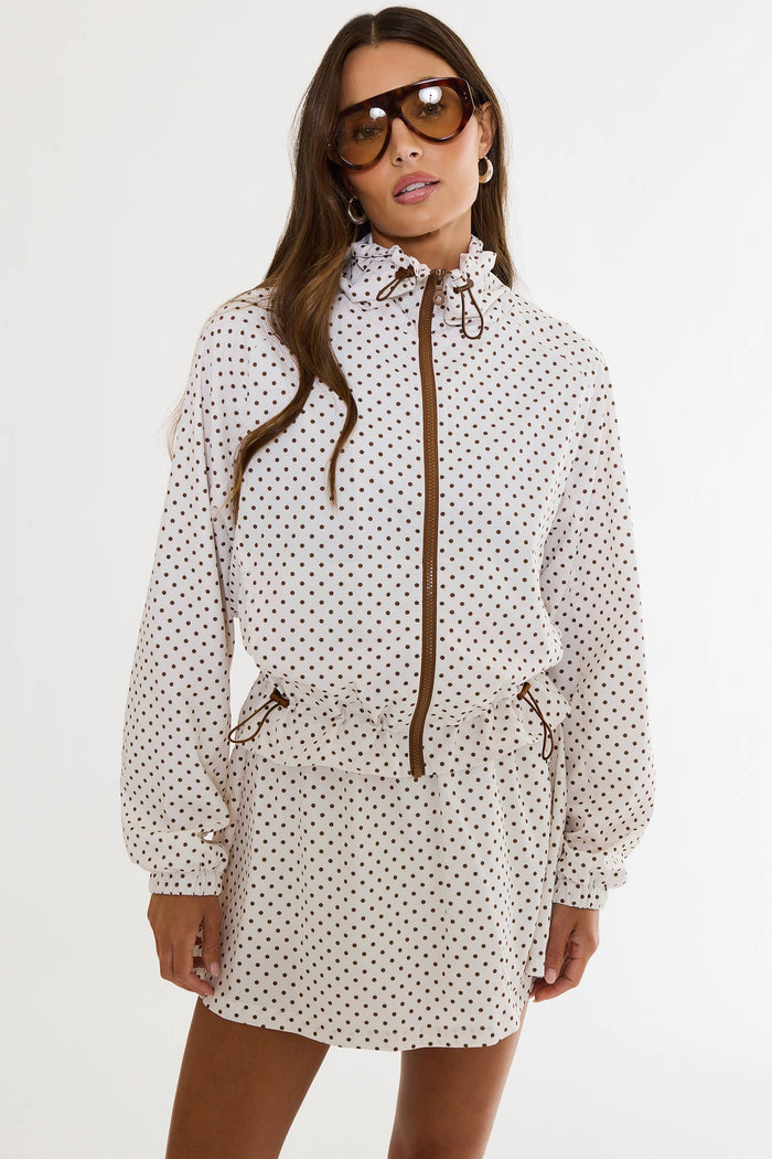 A woman modeling a white hooded raincoat dress with a brown polka dot pattern, featuring a brown front zipper, drawstring hood, and side pockets, set against a plain white studio background.