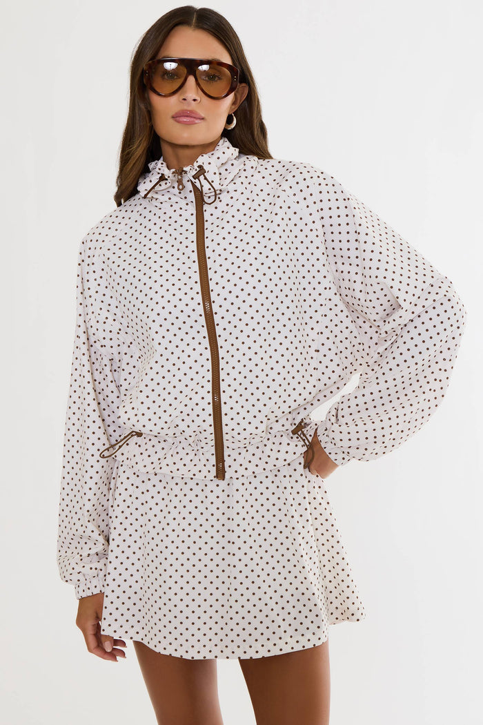 A woman modeling a white long-sleeve mini dress with a brown polka dot pattern, featuring a brown front zipper, drawstring collar, and matching drawstring waist, set against a plain white background.
