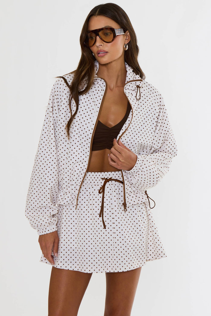 A woman modeling a matching white two-piece set with small red polka dots, featuring a zip-up jacket and a high-waisted mini skirt, against a plain white studio background.