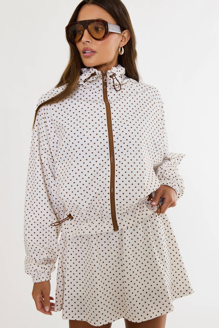 A woman wearing a white long-sleeve mini dress with a brown polka dot pattern, a brown zipper, and a high collar, styled with brown sunglasses.