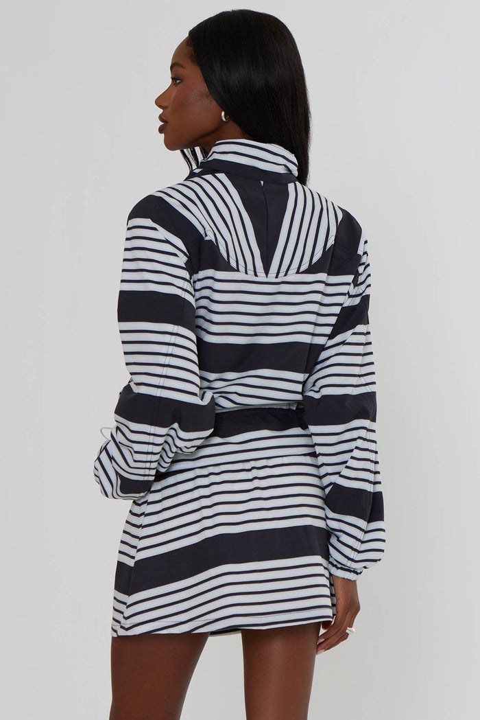 A model is shown from the back wearing a black and white horizontally striped long-sleeve zip-up jacket dress, paired with white socks and black and white sneakers, against a plain light gray studio background.