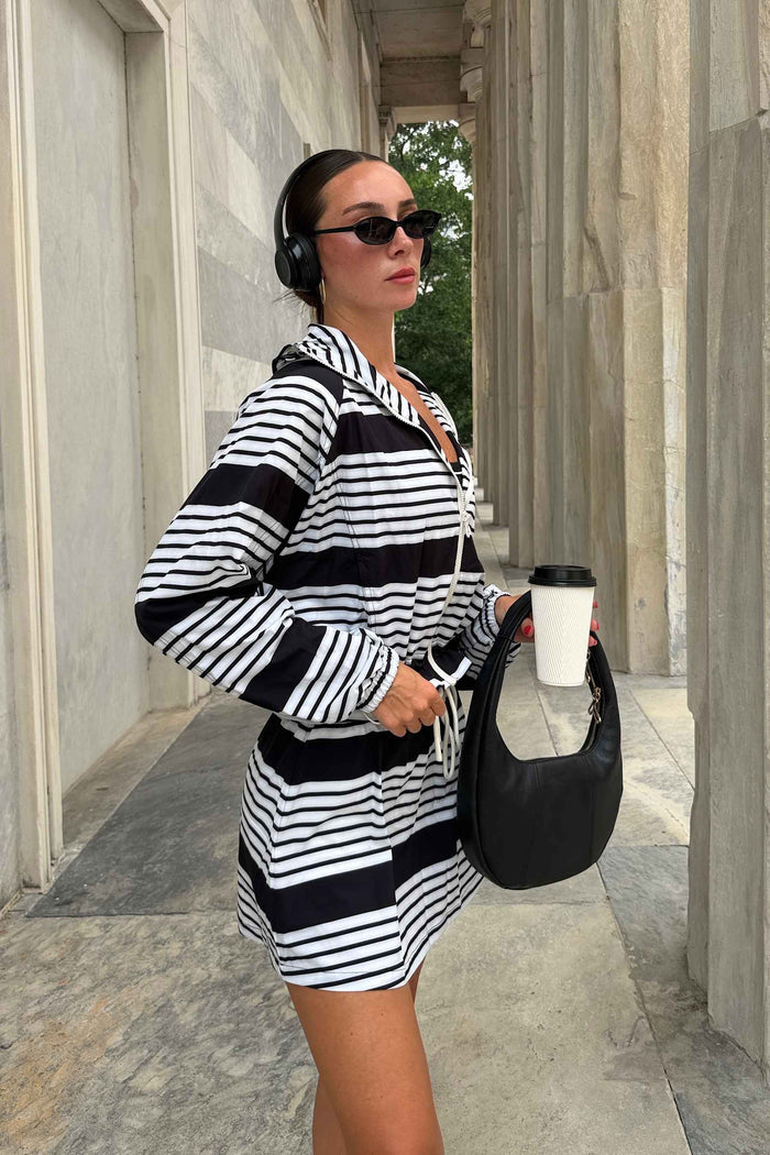 A woman wearing a black and white horizontally striped hooded sweatshirt dress stands in a stone colonnade, holding a white coffee cup and a black crescent shoulder bag.