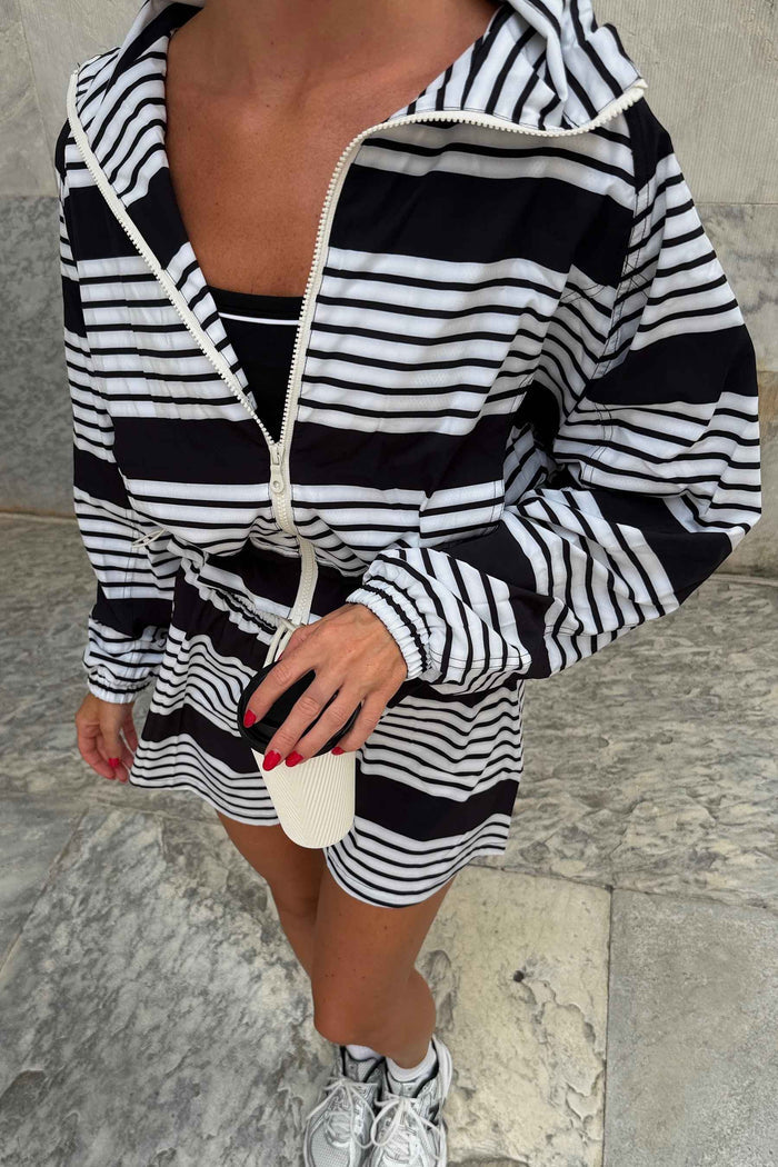 A person wearing a black and white horizontally striped zip-up hoodie and matching striped shorts, holding a small white handbag, standing on a stone surface.