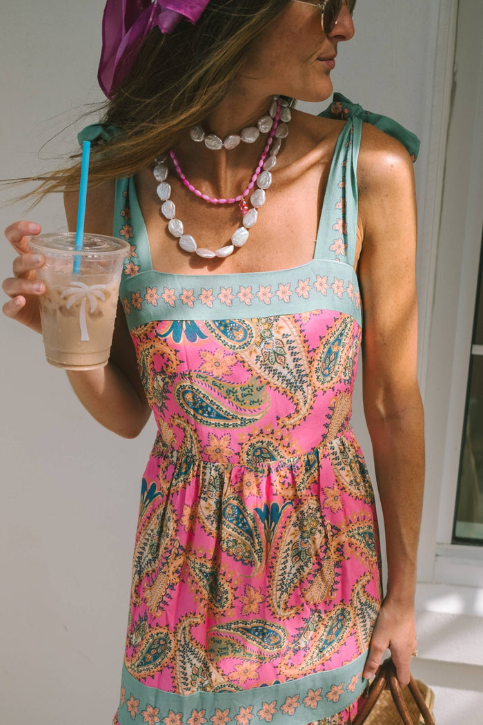 A woman modeling a sleeveless tiered maxi dress with a vibrant pink paisley pattern and turquoise green trim