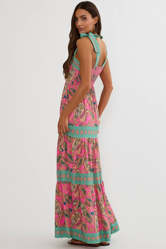 A woman modeling a sleeveless tiered maxi dress with a vibrant pink paisley pattern and turquoise green trim, shown from a three-quarters back view against a light gray studio background.