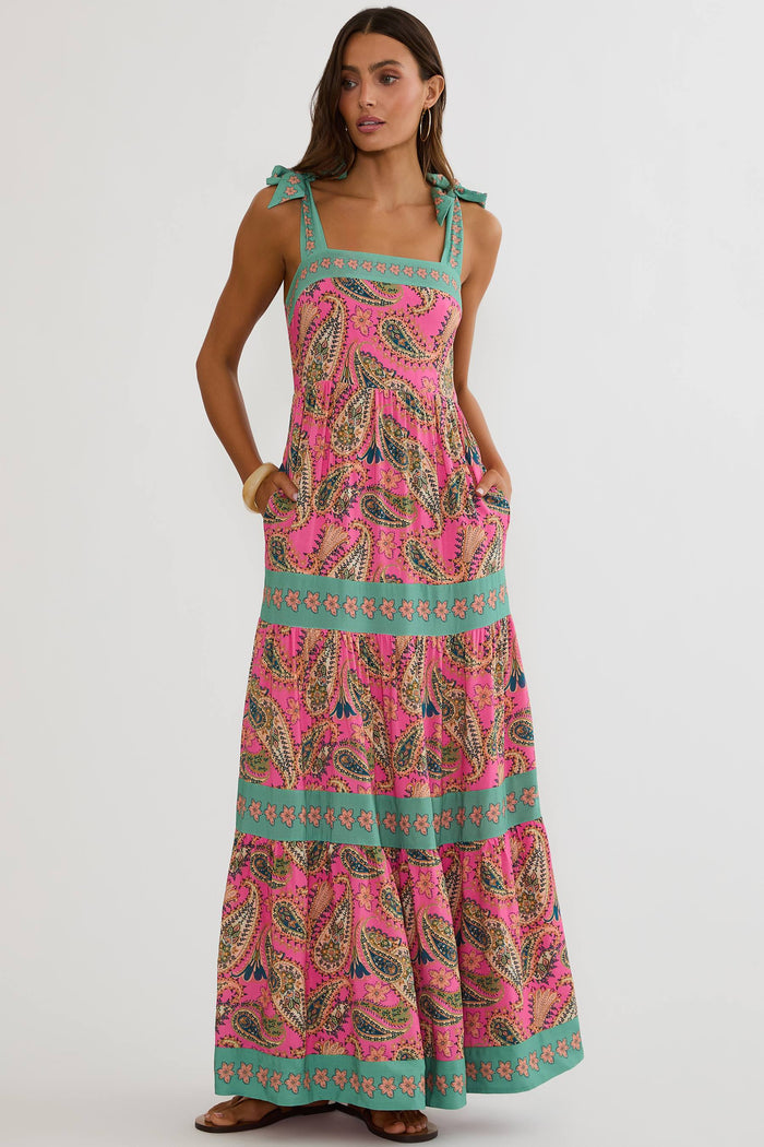 A woman modeling a pink sleeveless maxi dress with a teal and brown paisley pattern, tie shoulder straps, and side pockets, set against a plain light gray studio background.