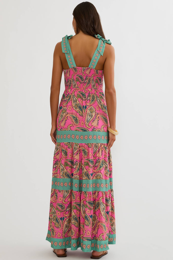 A woman viewed from the back wearing a sleeveless tiered maxi dress with a bright pink paisley print and turquoise green trim, against a plain light gray studio background.