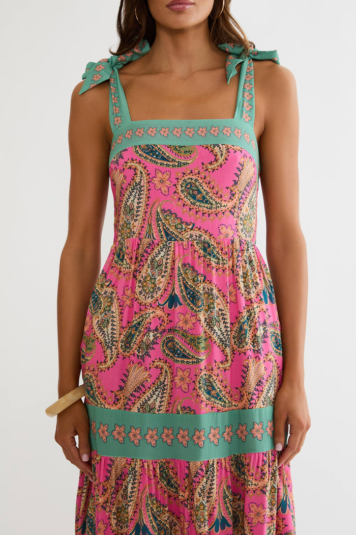 A woman modeling a sleeveless tiered maxi dress with a vibrant pink paisley pattern and turquoise green trim, shown from a three-quarters back view against a light gray studio background.