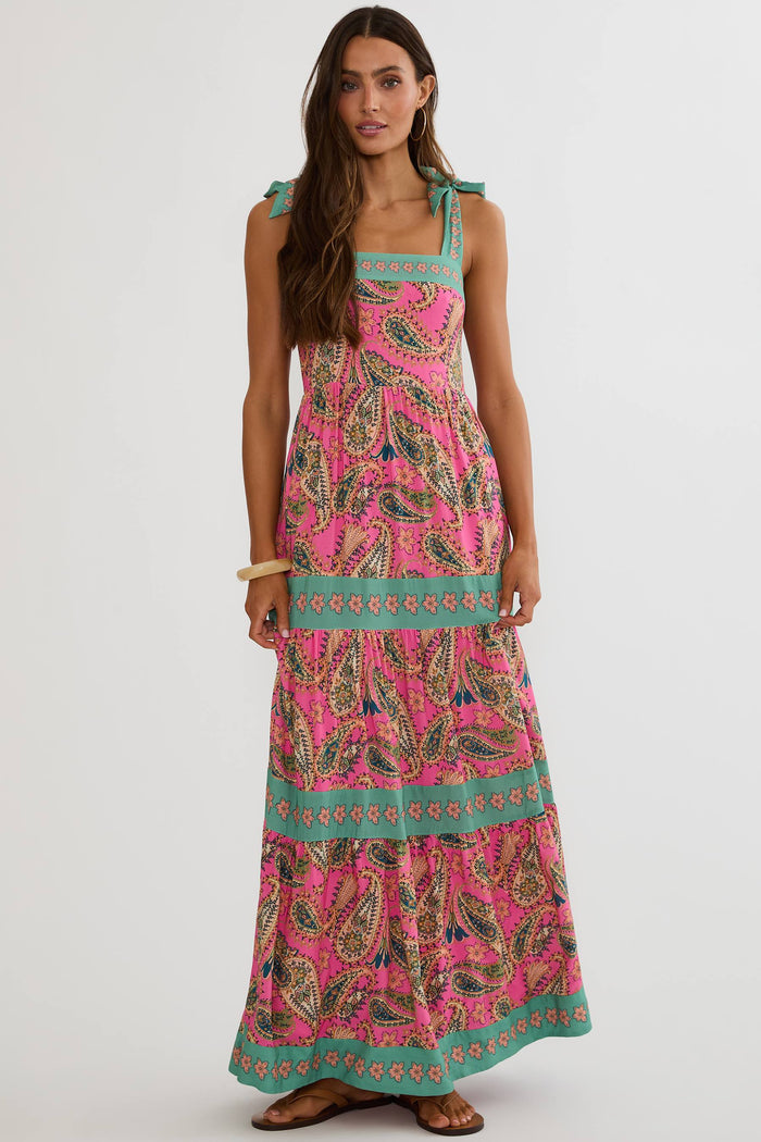 A woman modeling a sleeveless tiered maxi dress with a vibrant pink paisley pattern and teal green trim, set against a plain light gray studio background.