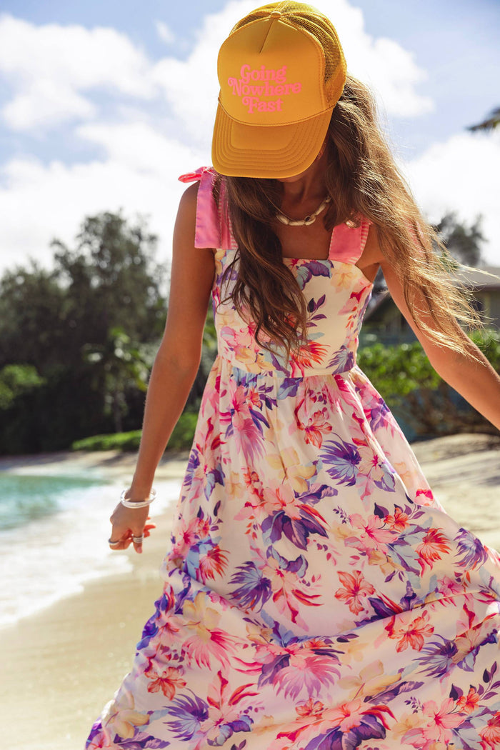Woman in a floral dress and yellow cap with pink text on a beach.