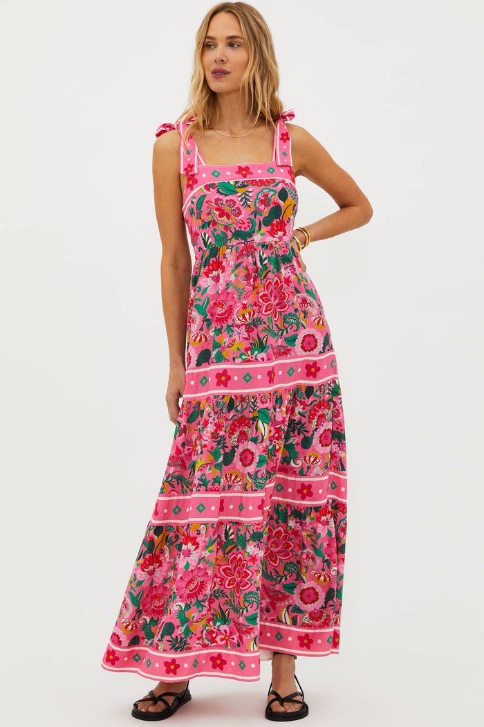 A model wearing a sleeveless, square neck, maxi dress in a vivid pink floral print with bow ties at the shoulders.