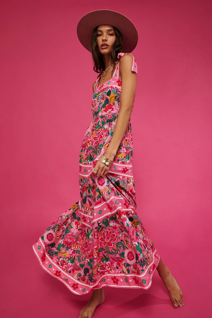 A model wearing a sleeveless, square neck, maxi dress in a vivid pink floral print with bow ties at the shoulders.