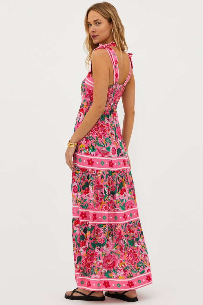 A model wearing a sleeveless, square neck, maxi dress in a vivid pink floral print with bow ties at the shoulders.