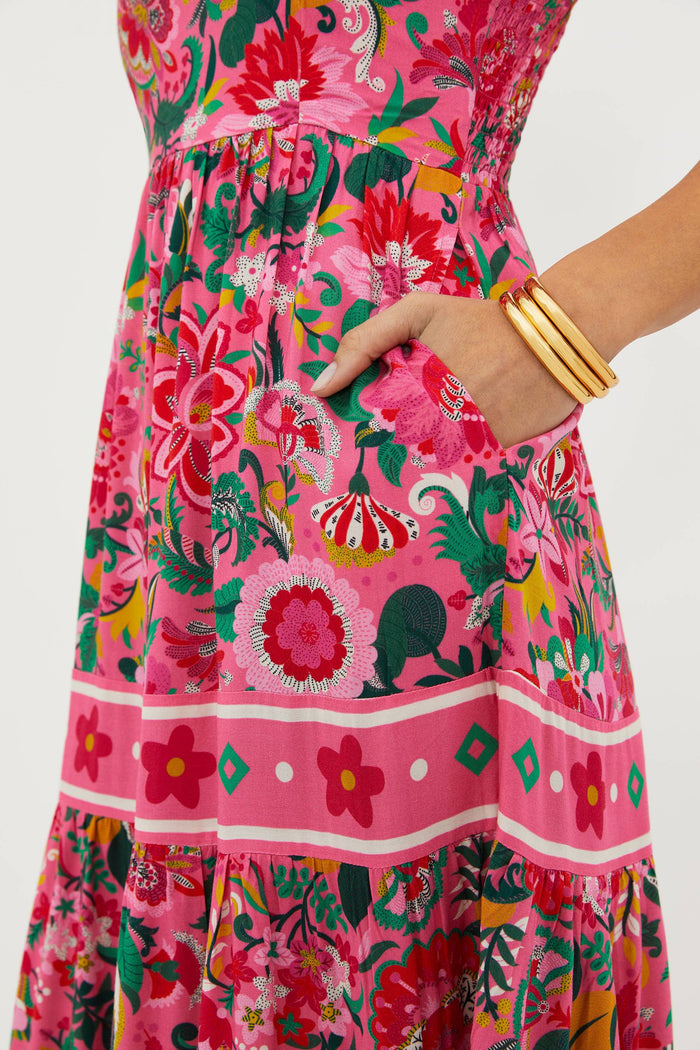 A model wearing a sleeveless, square neck, maxi dress in a vivid pink floral print with bow ties at the shoulders.