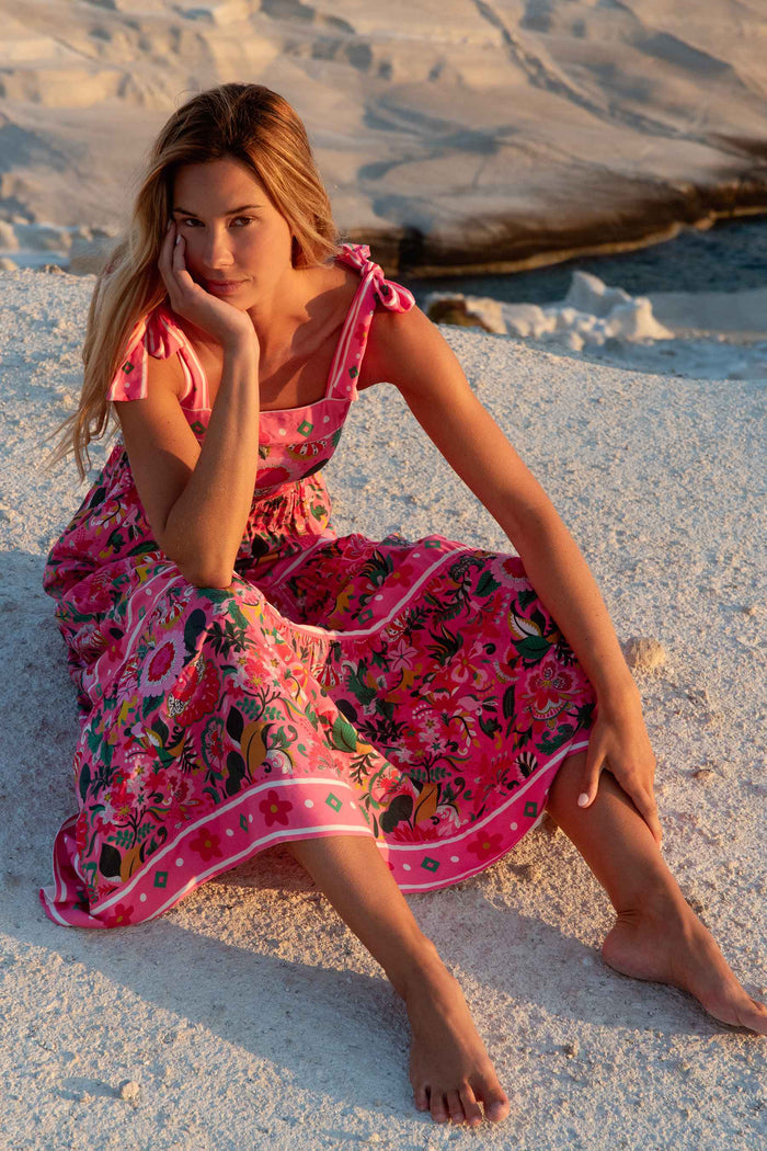 A model wearing a sleeveless, square neck, maxi dress in a vivid pink floral print with bow ties at the shoulders.