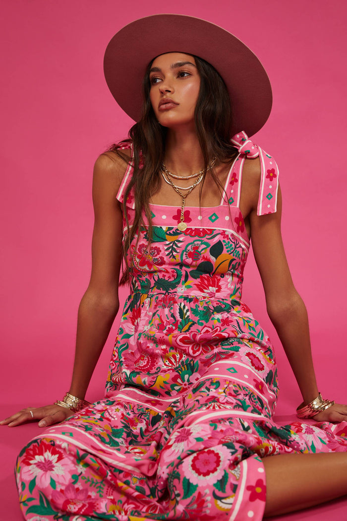A model wearing a sleeveless, square neck, maxi dress in a vivid pink floral print with bow ties at the shoulders.