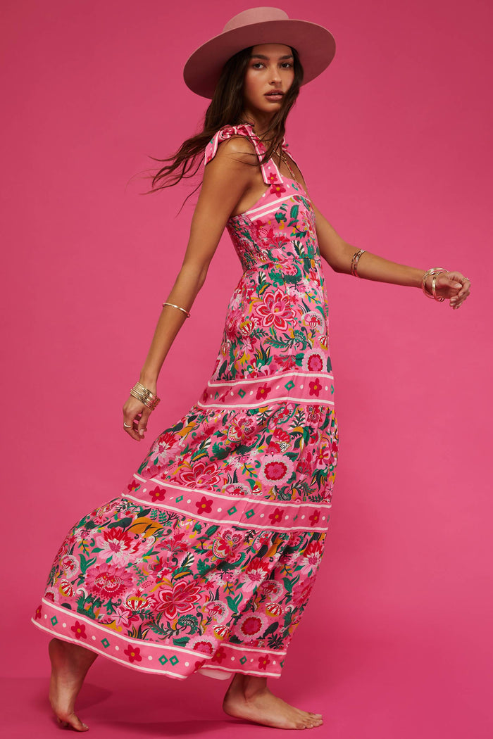 A model wearing a sleeveless, square neck, maxi dress in a vivid pink floral print with bow ties at the shoulders.