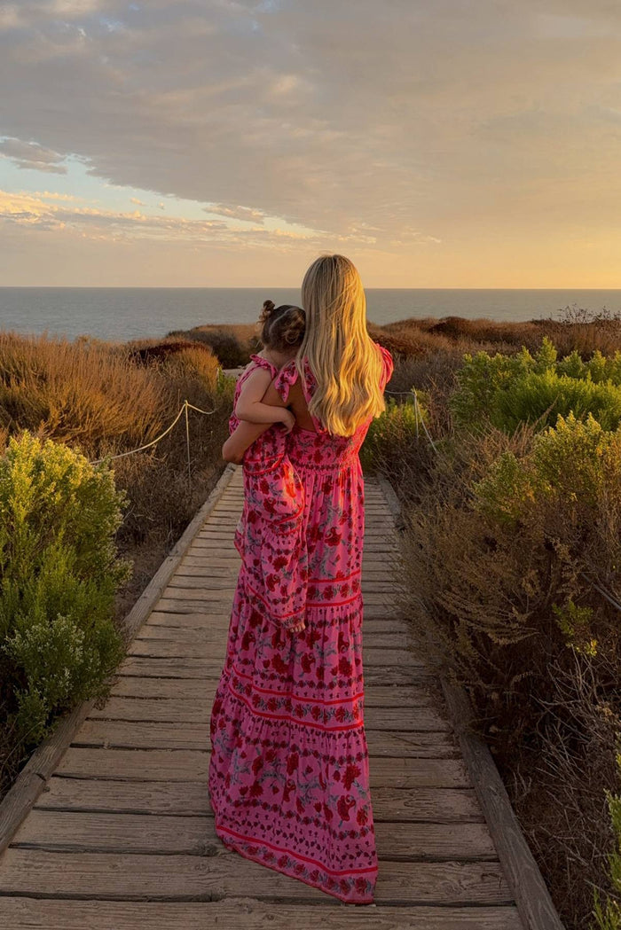 Woman in a pink dress holding a child on a wooden path at sunset.