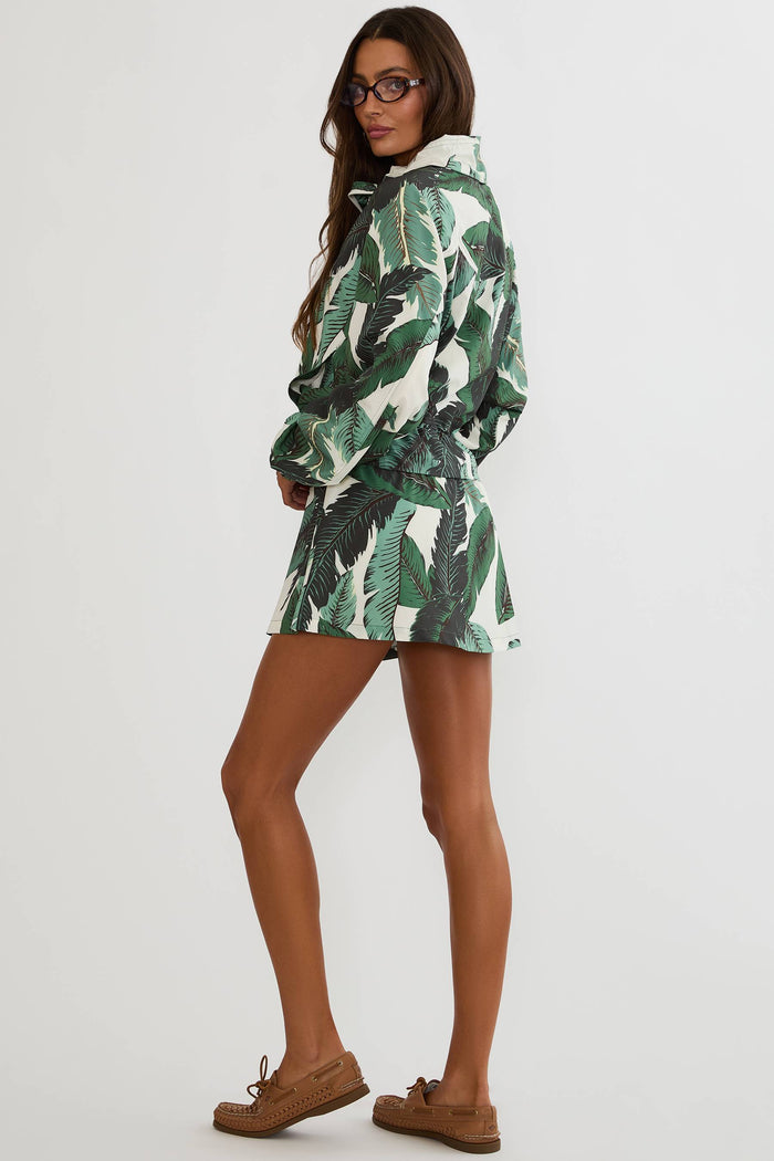 A woman modeling a white hooded raincoat with a green tropical leaf print, paired with brown boat shoes, against a plain light gray background.