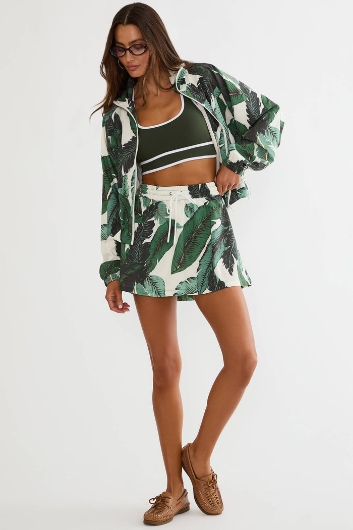 A woman modeling a matching tropical leaf print set consisting of a cropped tank top with white stripes, a matching zip-up jacket, and a high-waisted mini skirt, paired with brown boat shoes against a white studio background.