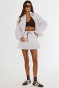 A model wearing a matching white and black polka dot lounge set consisting of a cropped long-sleeve shirt and a mini skirt with a drawstring waist, styled with a brown crop top, brown socks, and black-and-white sneakers against a plain white studio background.