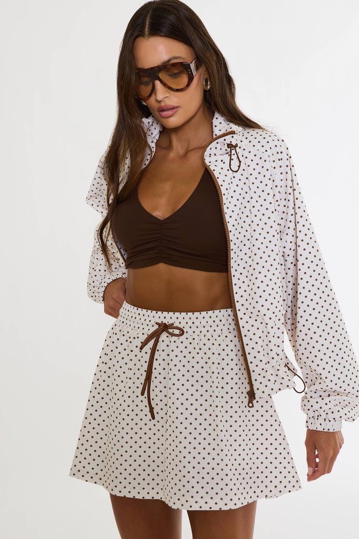 A woman modeling a matching two-piece set consisting of a brown ruched crop top and a white skirt with small black polka dots and a brown drawstring, styled with a matching white polka dot zip-up jacket and sunglasses.
