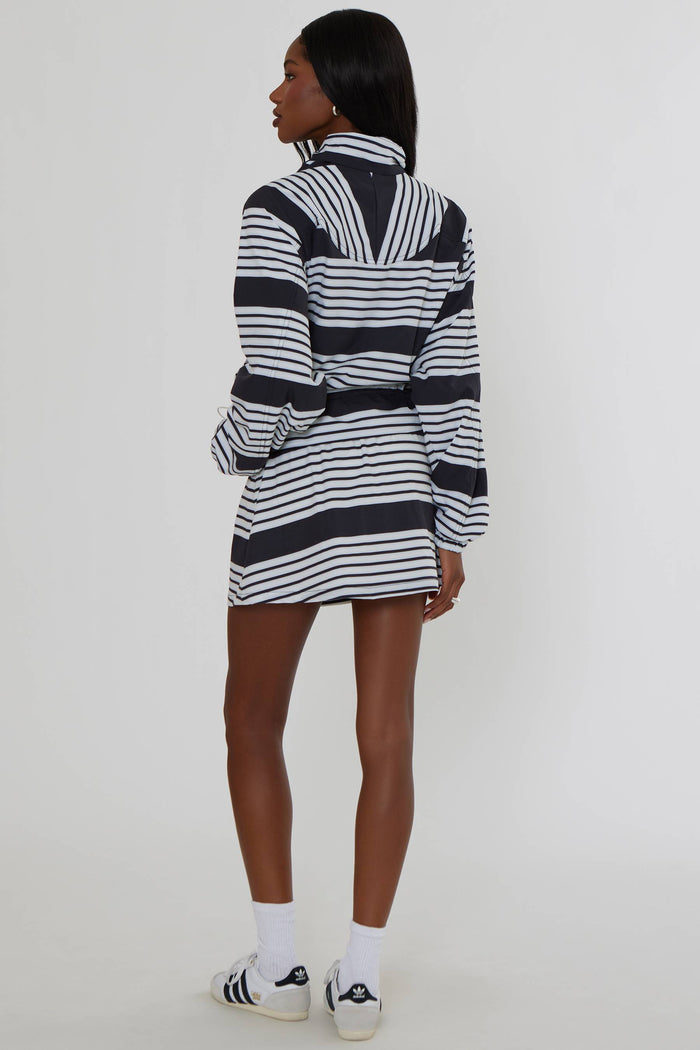 A model is shown from the back wearing a black and white horizontally striped long-sleeve zip-up jacket dress, paired with white socks and black and white sneakers, against a plain light gray studio background.