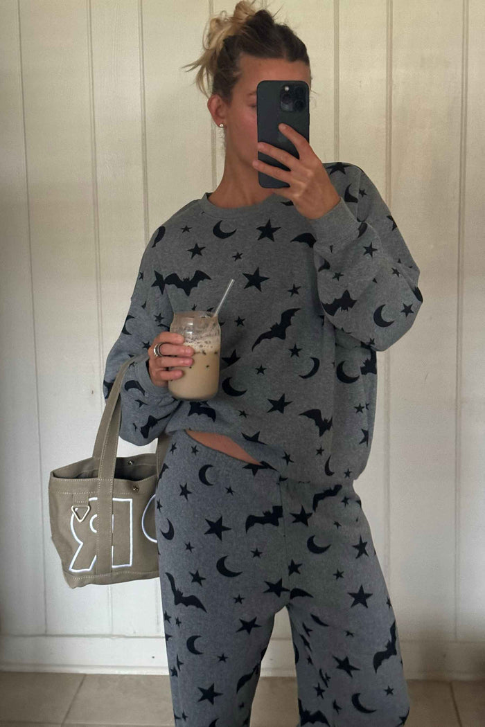 A woman takes a mirror selfie wearing a gray matching lounge set with a black moon and star pattern. She is holding an iced coffee in a clear cup and carrying a beige tote bag with a white graphic, set against a white paneled wall.