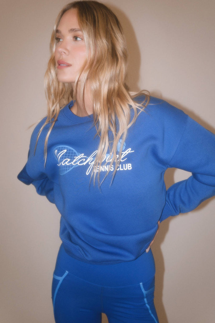A woman wearing a royal blue sweatshirt and matching leggings with 'Catch it Tennis Club' printed in white across the chest, posed against a plain beige wall.