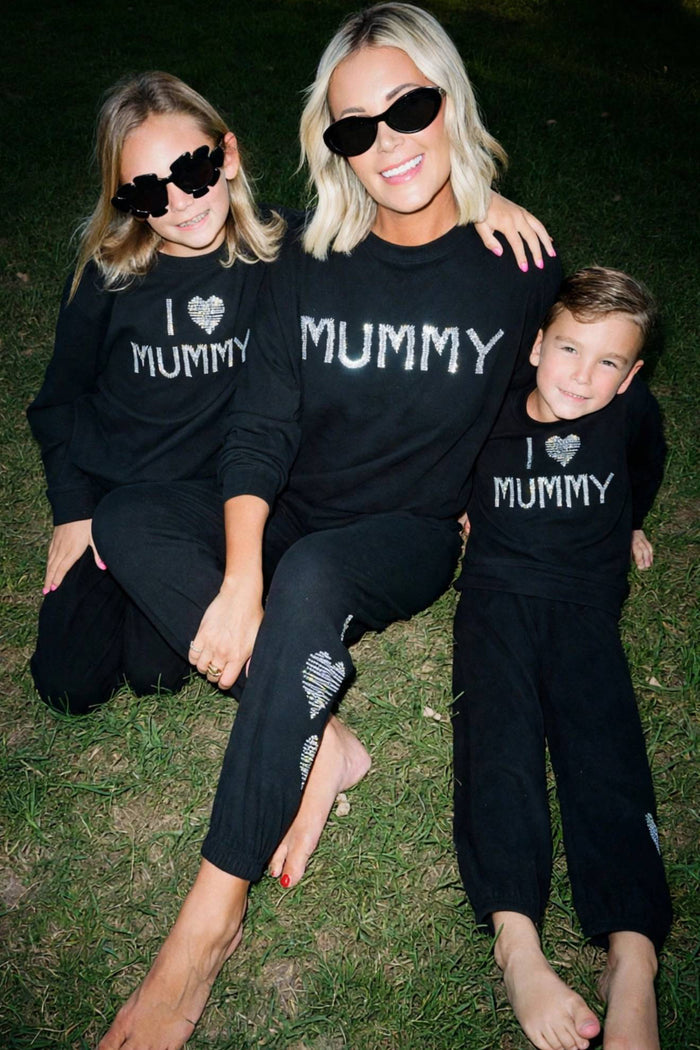 A woman and two children wearing matching black long-sleeve sweatshirts with white text reading 'I [heart] MUMMY' and 'MUMMY' sit together on grass at night.