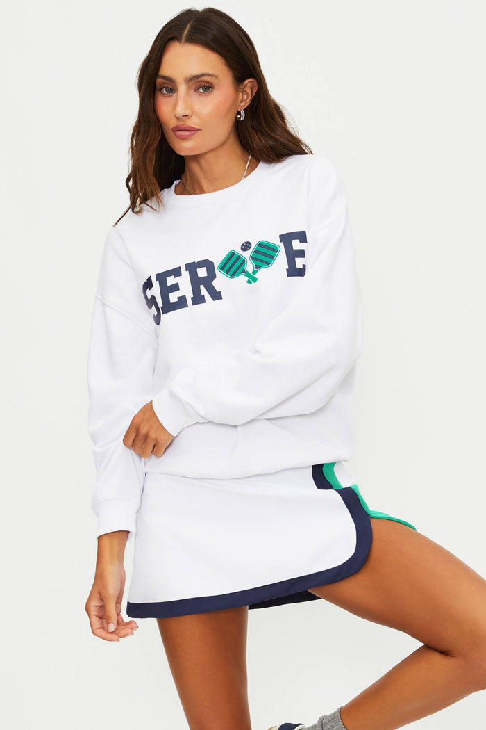 Woman wearing a white sweatshirt with 'SERVE' printed on it and a skirt with green and blue accents.