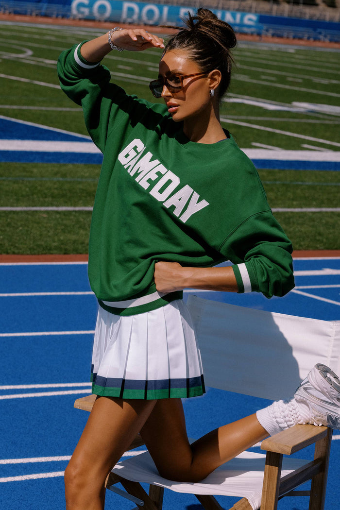 Person wearing a green 'GIMME DAY' sweatshirt and white skirt on a sports field.