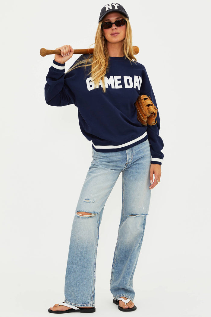 Person wearing a navy 'GAMEDAY' sweatshirt, light blue jeans, and a baseball cap with a bat and glove.