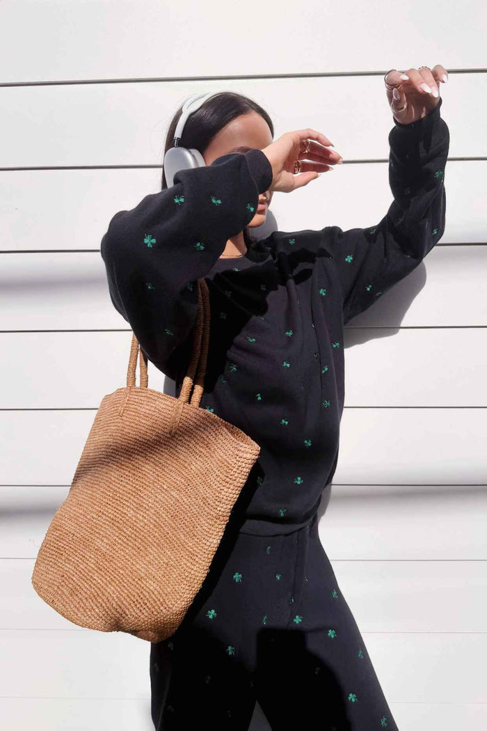 Person wearing a black hoodie with green patterns and holding a woven bag against a white background