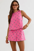 A woman modeling a bright pink sleeveless top and matching shorts set with a small blue pattern, styled with sunglasses, gold bracelets, and brown sandals against a light gray studio background.