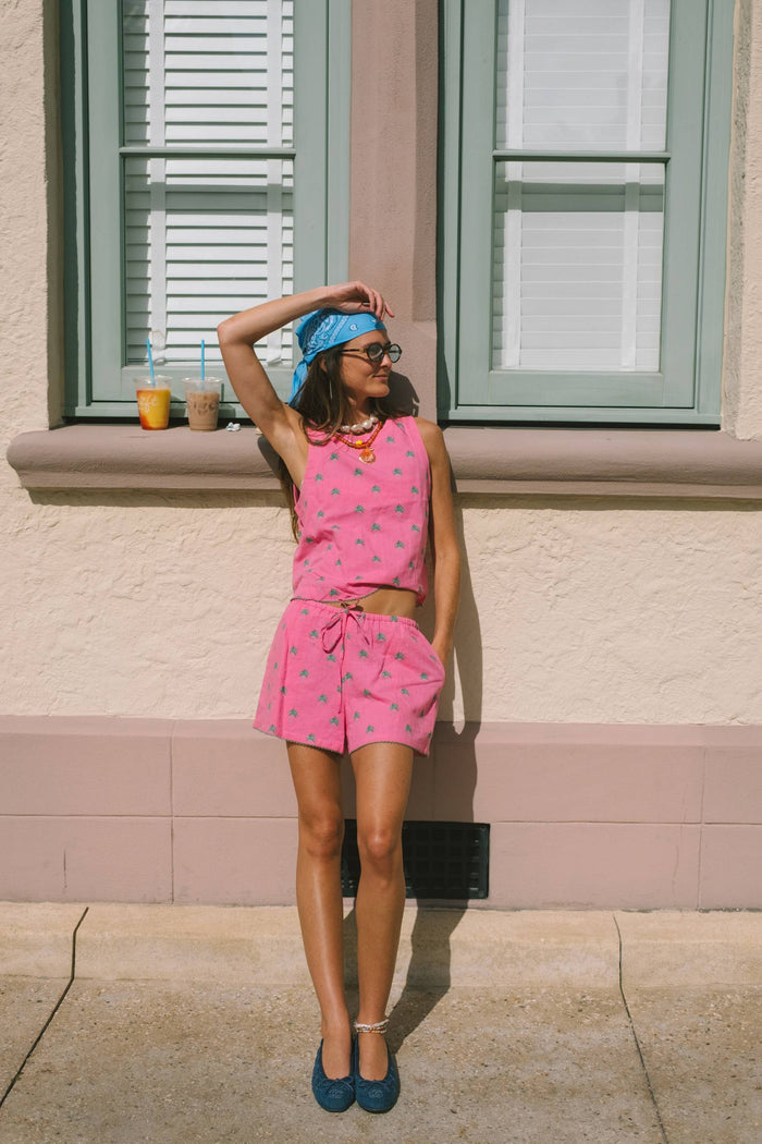 A woman stands outdoors against a beige stucco building wall with green shutters, wearing a bright pink two-piece outfit with a small repeating pattern, a blue headscarf, and blue shoes.