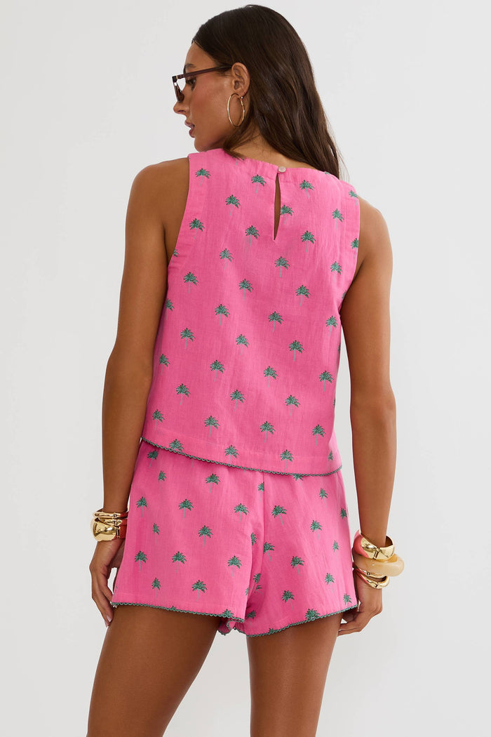 A woman modeling a bright pink sleeveless top and matching shorts set with a small blue pattern, styled with sunglasses, gold bracelets, and brown sandals against a light gray studio background.