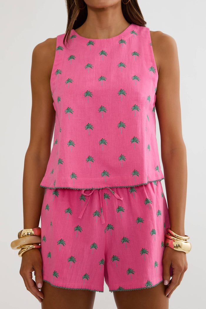 A woman wearing a bright pink sleeveless top and matching shorts set with a small green palm tree pattern, photographed against a light gray studio background.