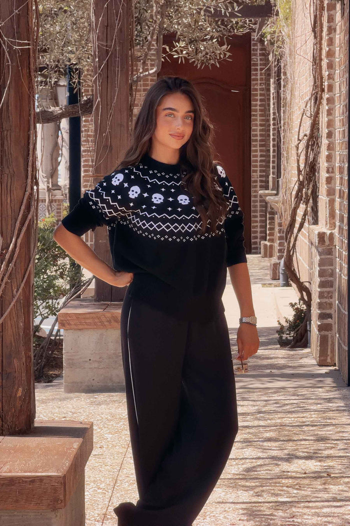 A woman stands in a sunlit outdoor alley wearing a black knit top with a white geometric and floral pattern across the chest and shoulders, paired with black wide-leg pants.