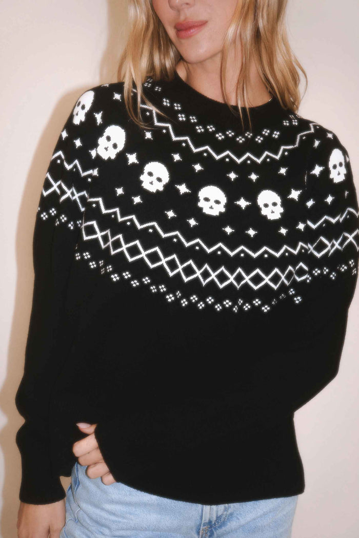A woman wearing a black knit sweater with a white Nordic-style pattern featuring skulls, stars, and geometric shapes, paired with light blue jeans.