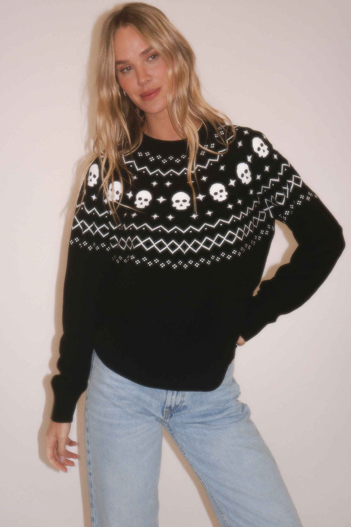 A woman modeling a black knit sweater with a white Nordic-style pattern featuring skulls, stars, and geometric designs across the chest and shoulders, paired with light blue jeans against a plain light background.